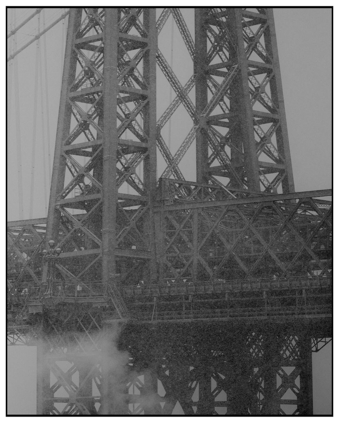 Monochrome shot of a towering bridge structure featuring a complex network of steel beams against a cloudy, overcast sky and wisps of smoke below.