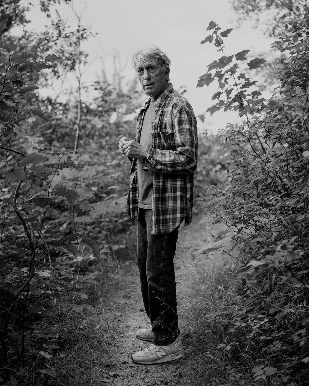 Black and white portrait of a mature man in a plaid shirt standing in a wooded area with trees and foliage surrounding him.