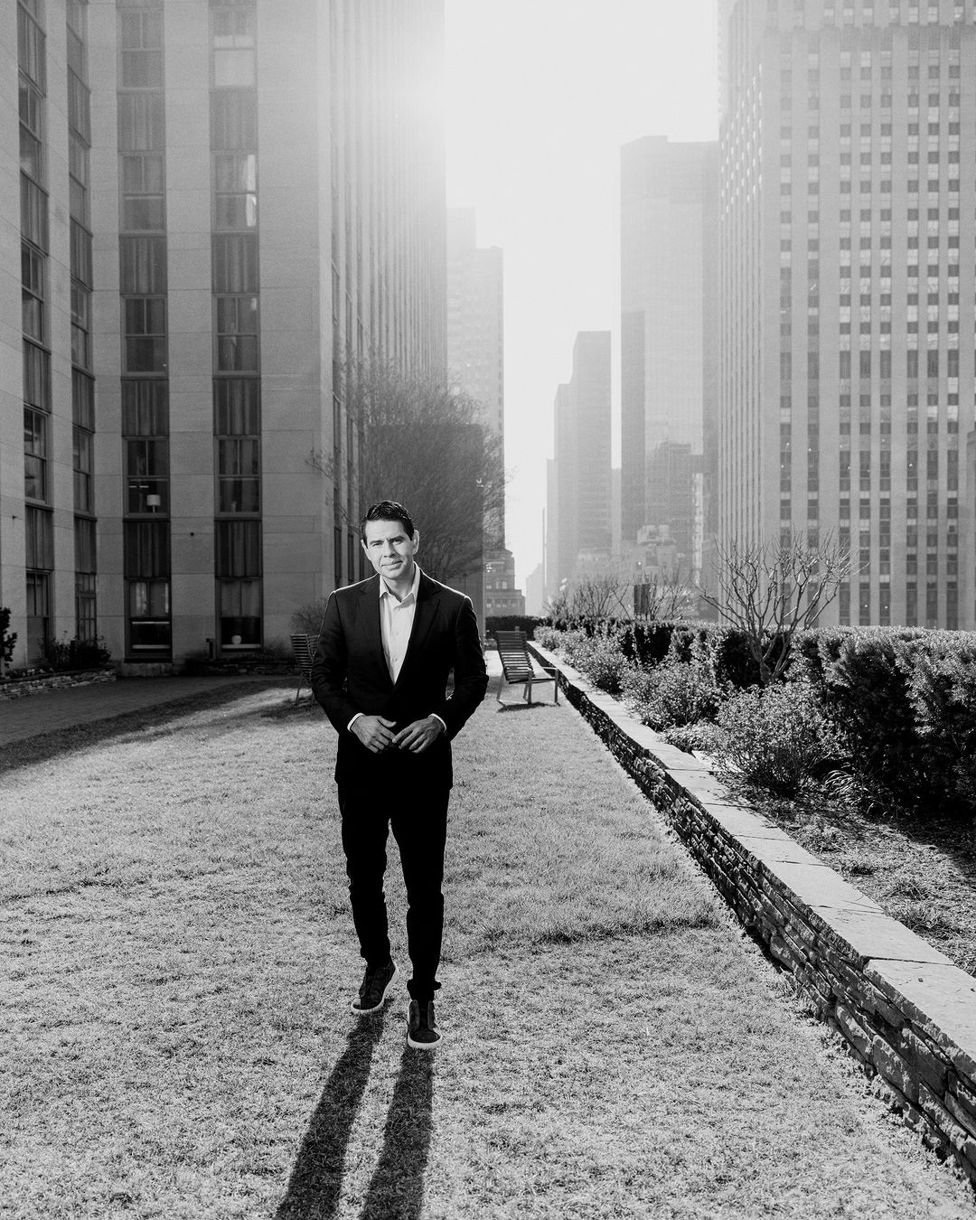 Adult male in a suit stands on a lawn in a city setting with skyscrapers and tall buildings on a sunny day. Black and white.
