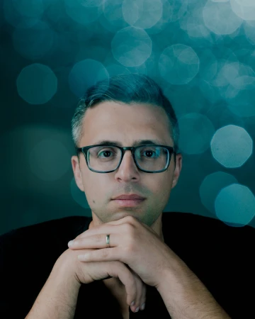 Portrait of a man in his 30s wearing glasses with hands under his chin against a blue bokeh background
