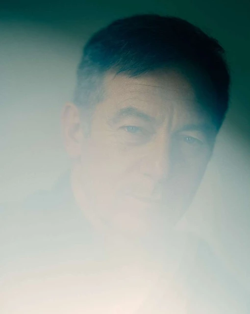 Portrait of an adult male with a blurred effect, wearing dark clothing, set against a soft gradient background with cool, neutral tones.