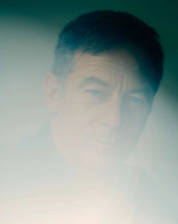 Portrait of an adult male with a blurred effect, wearing dark clothing, set against a soft gradient background with cool, neutral tones.