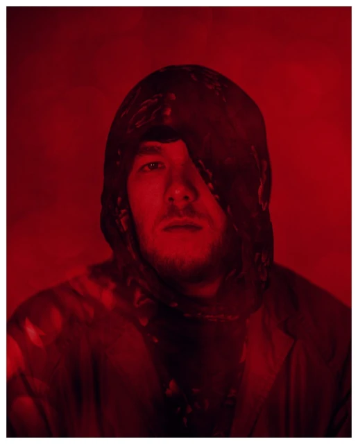 Portrait of a man with fair skin wearing a dark-colored hooded garment against a solid red background, creating a bold, monochromatic image.