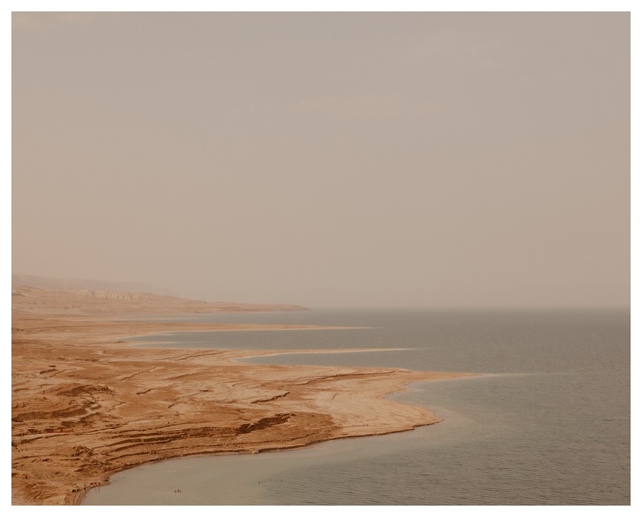 Aerial view of the Dead Sea shoreline with textured, sandy beaches meeting the calm, blue water under a hazy, overcast sky.