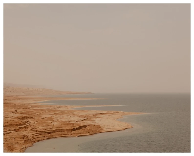 Aerial view of the Dead Sea shoreline with textured, sandy beaches meeting the calm, blue water under a hazy, overcast sky.