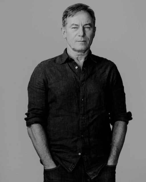 Black and white portrait of a mature adult man with hands in pockets, wearing a casual button-down shirt against a plain background.