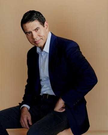 Man in a dark blazer and blue button-down shirt seated against a warm neutral background