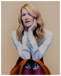 Laura Dern with blonde hair wearing a white shirt and brown skirt resting chin in hand against a neutral background