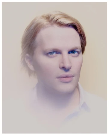 Close up portrait of a blonde man with blue eyes wearing a white shirt against a plain light background photographed by Benedict Evans