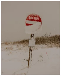 Snow-covered circular red do not enter sign mounted on a wooden post during a winter snowstorm.