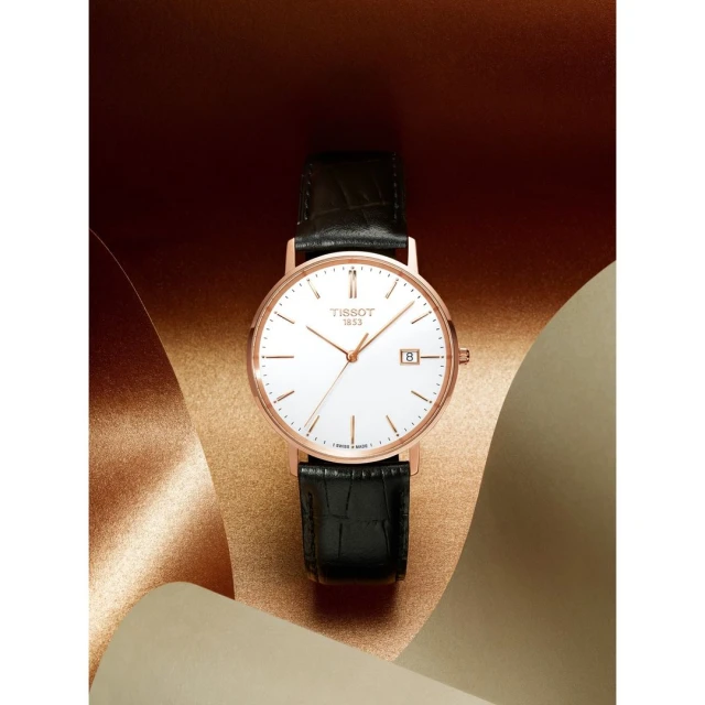 Rose gold Tissot watch with a white face and black leather strap on a tan surface, studio shot for a fashion or jewelry ad.