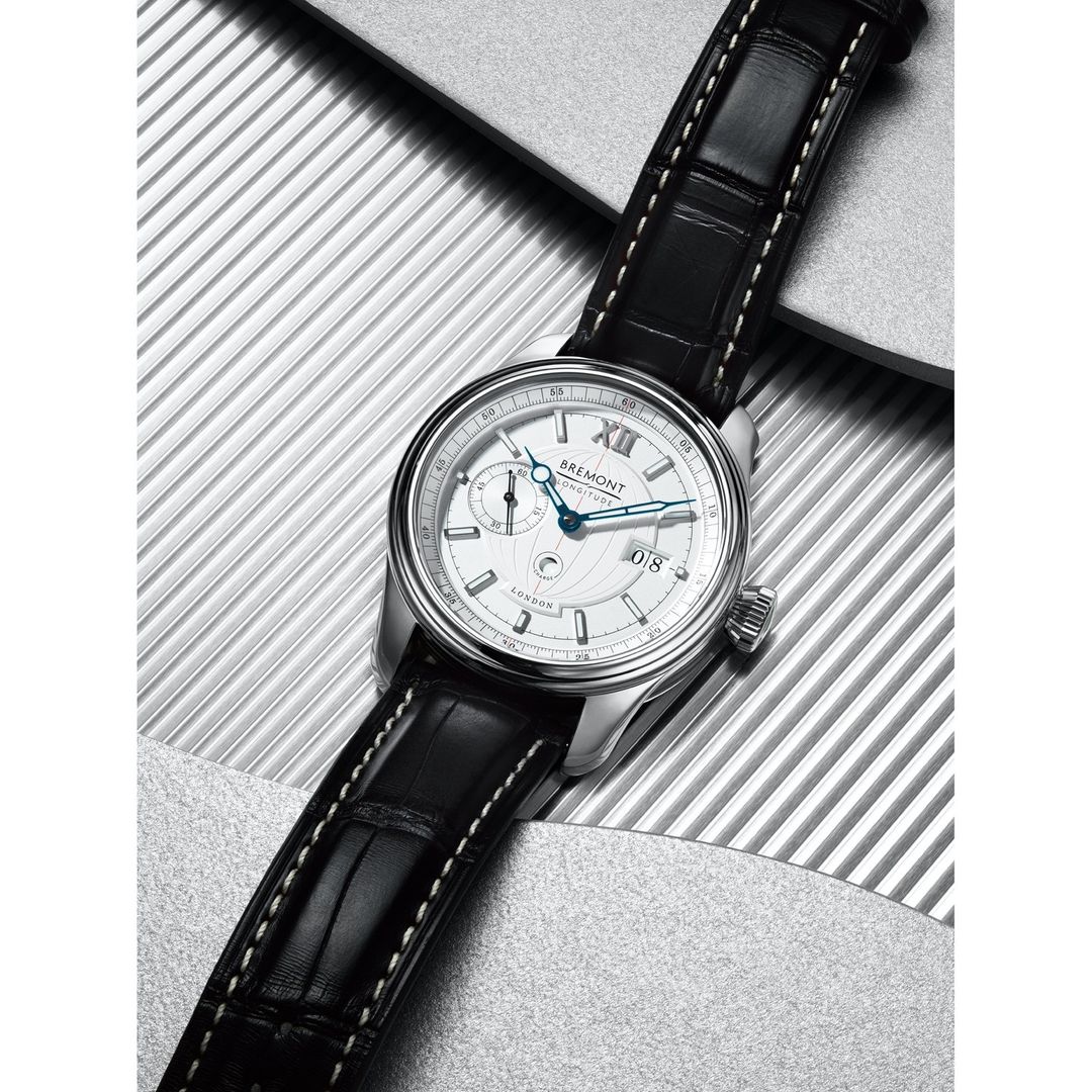 Silver Bremont wristwatch with a black leather strap, featuring a round white dial and a silver case, against a textured background.