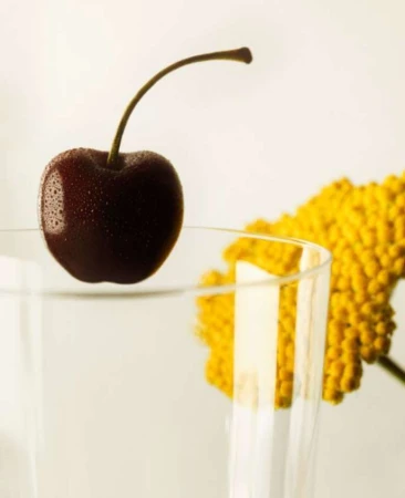 Close-up shot of a ripe cherry with water droplets on the surface, placed on the rim of a glass with a yellow flower in the background.
