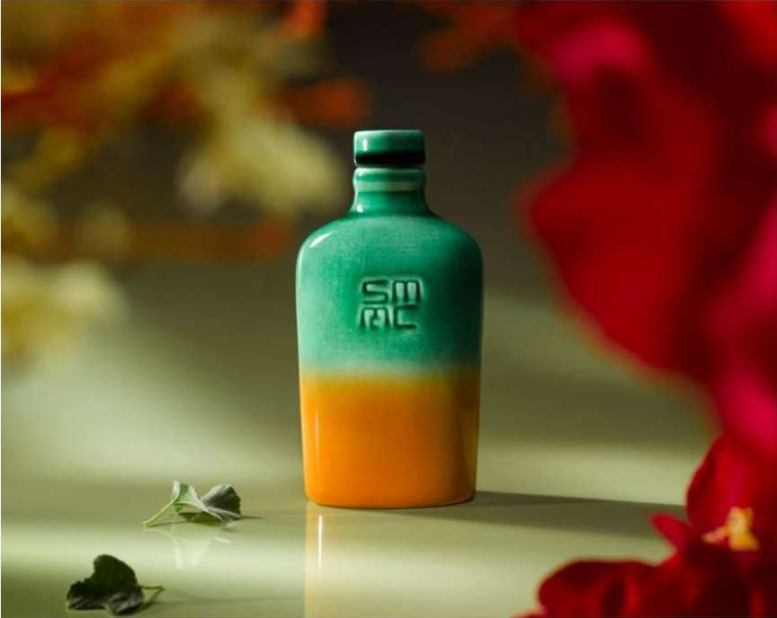 Two-tone green and orange ceramic bottle on a light green surface with flower petals creating a colorful blurred background.