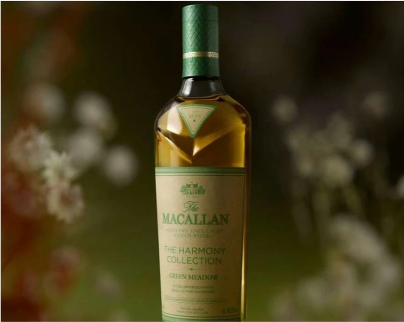A bottle of The Macallan Harmony Collection Green Meadow whisky stands centered against a blurred floral background, highlighting its green labeling.