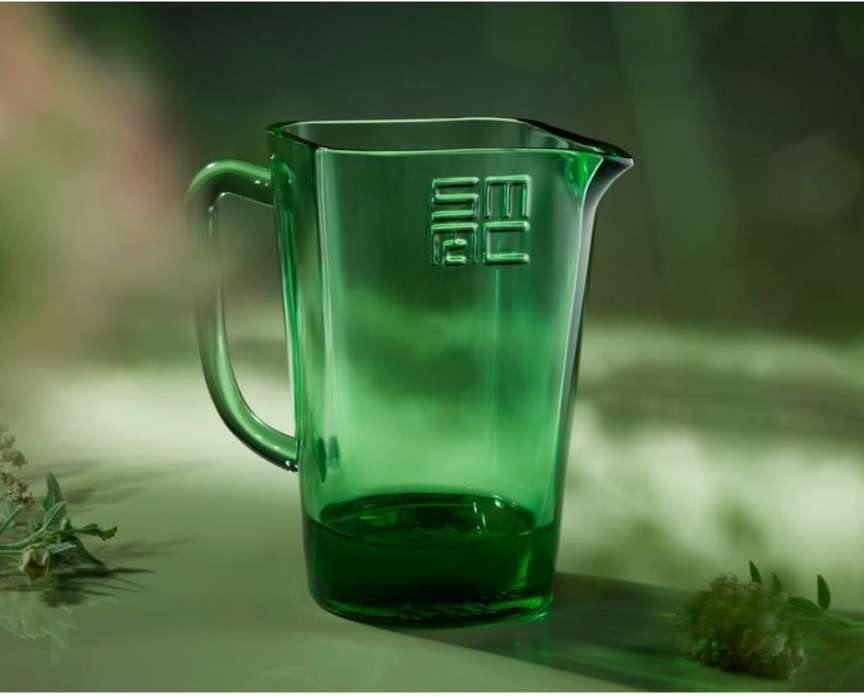 Close-up studio shot of a transparent green glass pitcher with an embossed logo, set against a soft green background with plant accents.