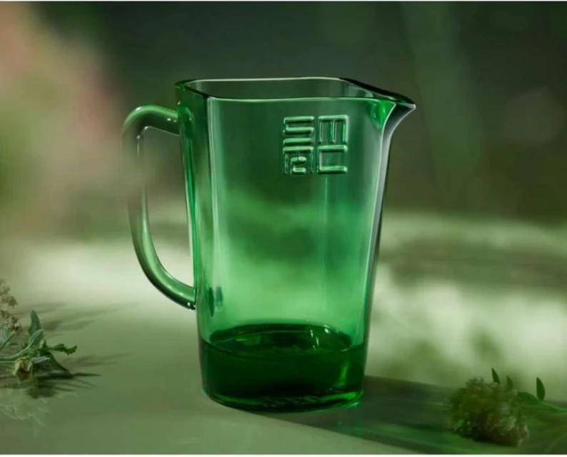 Close-up studio shot of a transparent green glass pitcher with an embossed logo, set against a soft green background with plant accents.