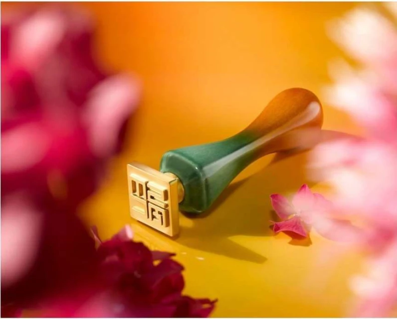 Close-up studio shot of a jade and gold stamp surrounded by blurred pink and red flowers against an orange and yellow background.