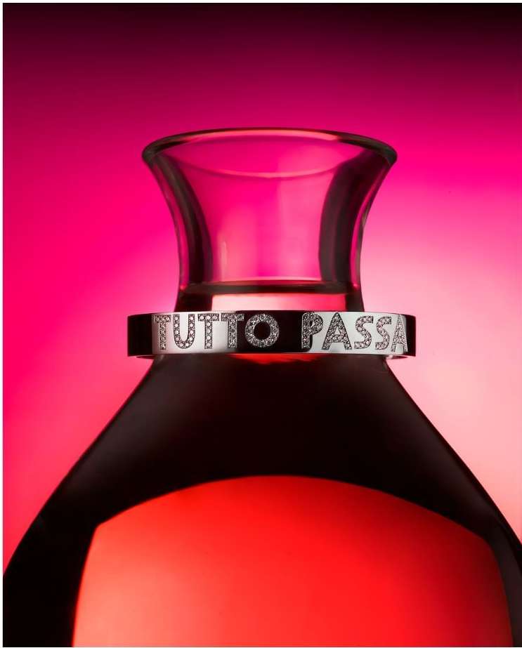 Elegant perfume bottle with clear glass stopper and silver band labeled 'TUTTO PASSA' set against a vibrant gradient pink background.