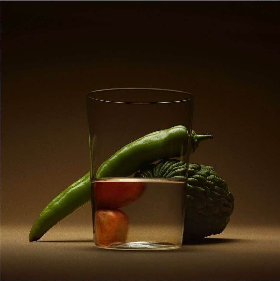 Still life featuring a clear glass filled with water, showcasing a green pepper, a red tomato, and a cherimoya, against a gradient brown backdrop.