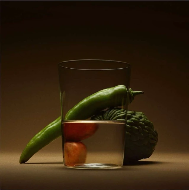 Still life featuring a clear glass filled with water, showcasing a green pepper, a red tomato, and a cherimoya, against a gradient brown backdrop.