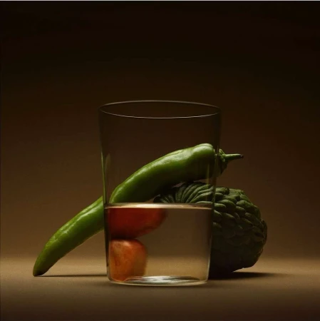 Still life featuring a clear glass filled with water, showcasing a green pepper, a red tomato, and a cherimoya, against a gradient brown backdrop.