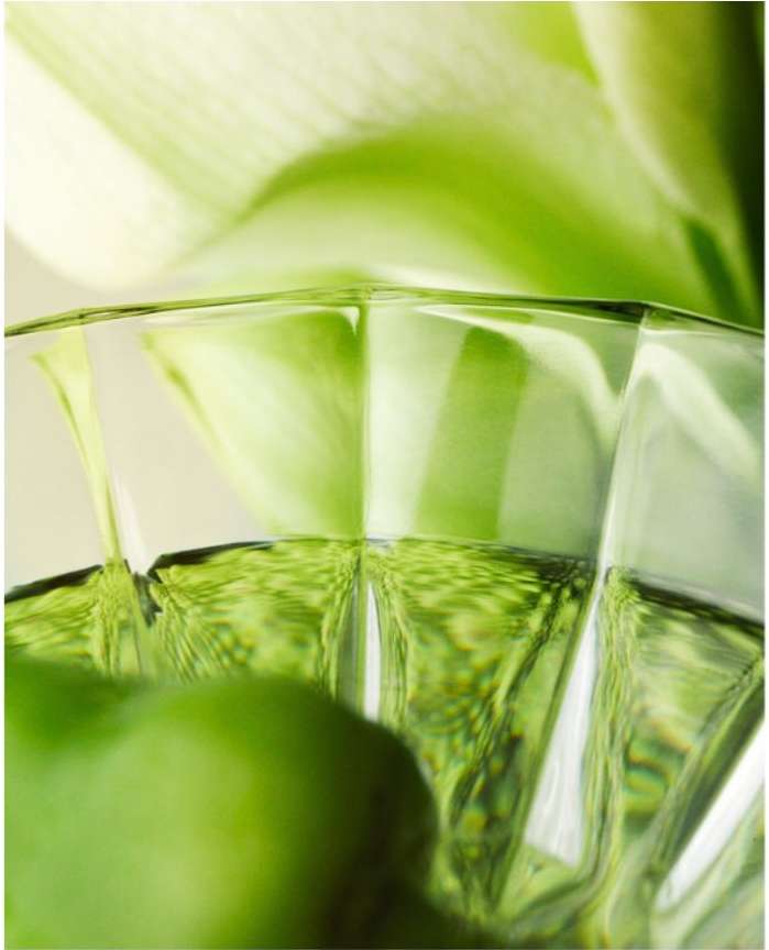 Close-up shot of a faceted clear glass filled with a light-green liquid, featuring a blurred background of a white Calla Lily.
