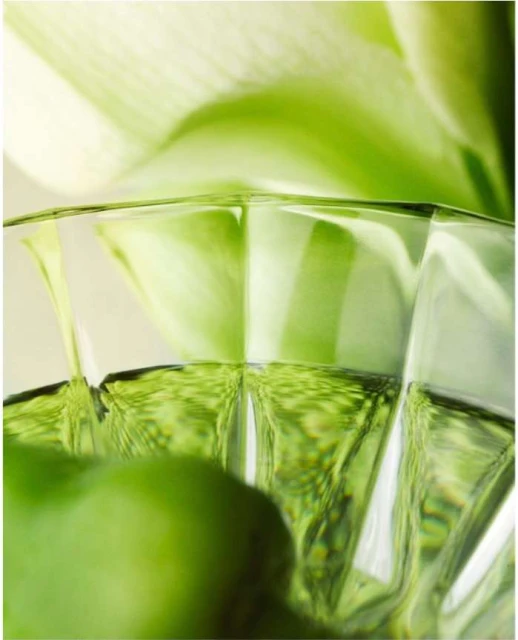 Close-up shot of a faceted clear glass filled with a light-green liquid, featuring a blurred background of a white Calla Lily.