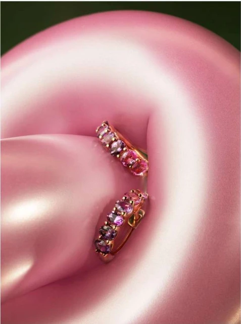 Close-up of a pair of gold hoop earrings with multiple pink and purple gemstones, set against a softly blurred pink background.
