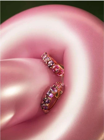 Close-up of a pair of gold hoop earrings with multiple pink and purple gemstones, set against a softly blurred pink background.