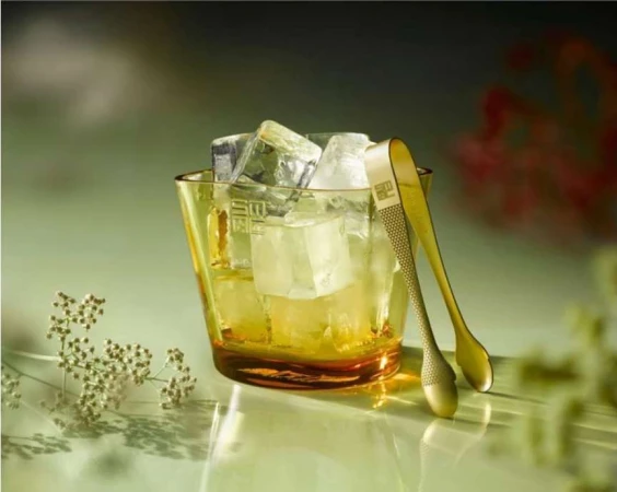 Ice cubes in a yellow glass bucket with gold tongs on a reflective surface with botanical accents and a soft, diffuse lighting.