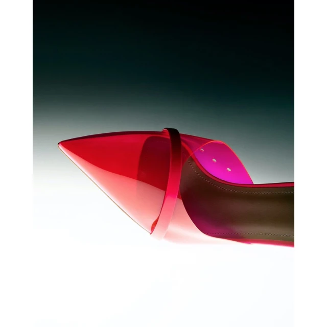 Elevated shot of a red, transparent, pointed-toe high heel shoe, showcasing the design and craftsmanship against a dark background.