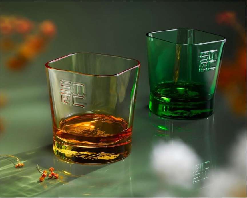 Two square-shaped glasses, one amber and one green, sit on a glossy green surface with sparse floral decorations, in a product shot.