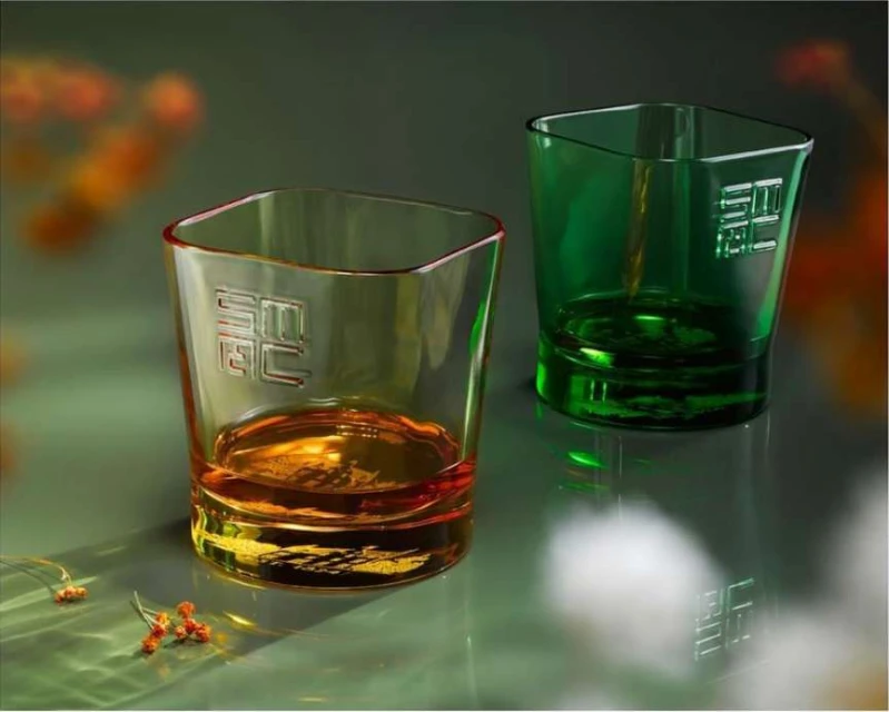 Two square-shaped glasses, one amber and one green, sit on a glossy green surface with sparse floral decorations, in a product shot.