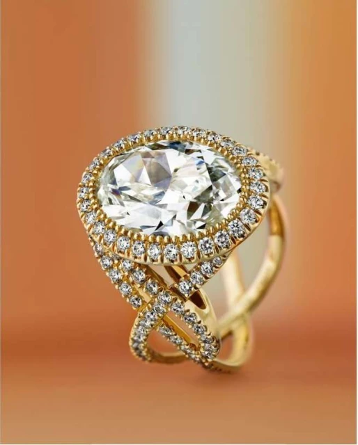 Close-up of a gold ring with a large oval diamond surrounded by smaller diamonds, showcasing jewelry design and fine craftsmanship on gradient backdrop.