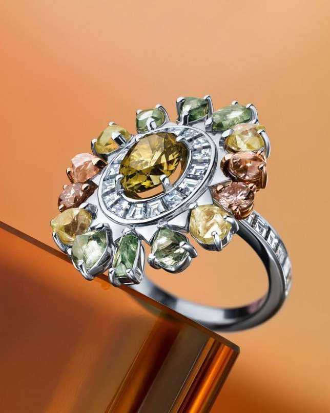 High-angle close-up of a ring made of white metal and multiple gemstones in various colors, set against a gradient orange background.