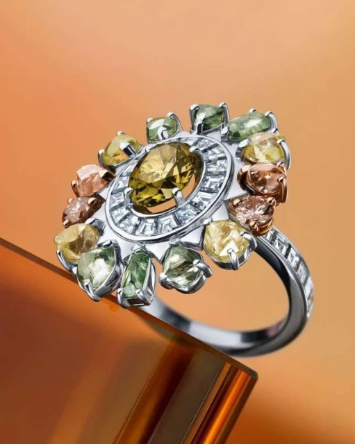 High-angle close-up of a ring made of white metal and multiple gemstones in various colors, set against a gradient orange background.