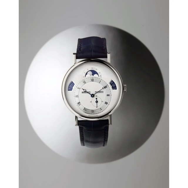 Silver and blue luxury wristwatch with a moon phase complication on a glossy, dappled grey surface, elevated for a product shot.