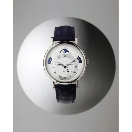 Silver and blue luxury wristwatch with a moon phase complication on a glossy, dappled grey surface, elevated for a product shot.