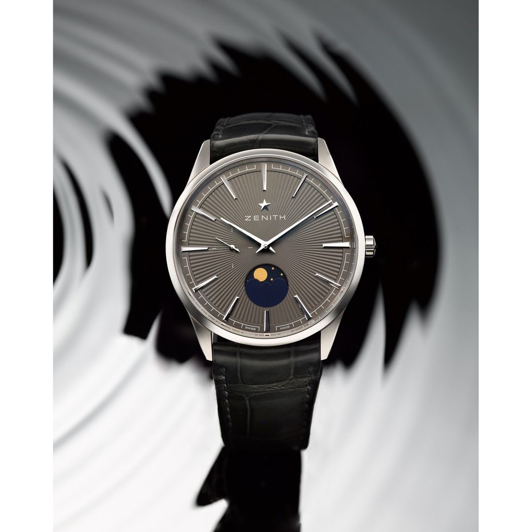 Elegant wristwatch featuring a gray dial with radial lines, moon phase indicator, and black leather strap on a neutral background.