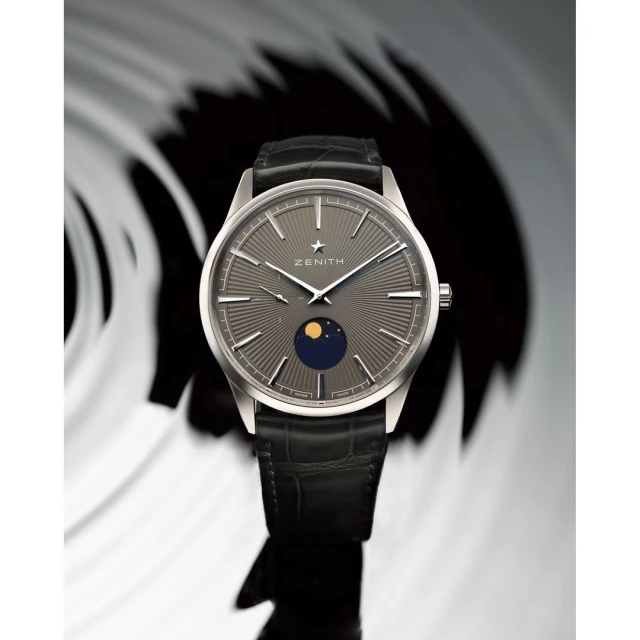 Elegant wristwatch featuring a gray dial with radial lines, moon phase indicator, and black leather strap on a neutral background.