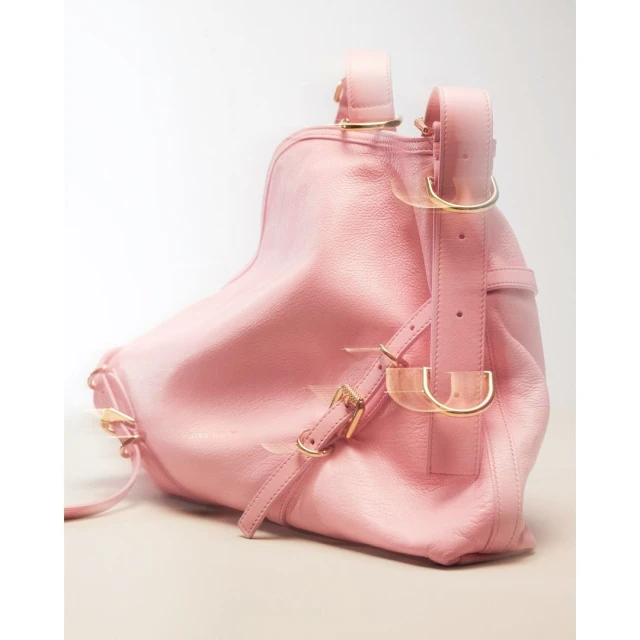 Close-up studio shot of a pink leather backpack with gold hardware, straps, and buckles against a white background, fashion accessory.