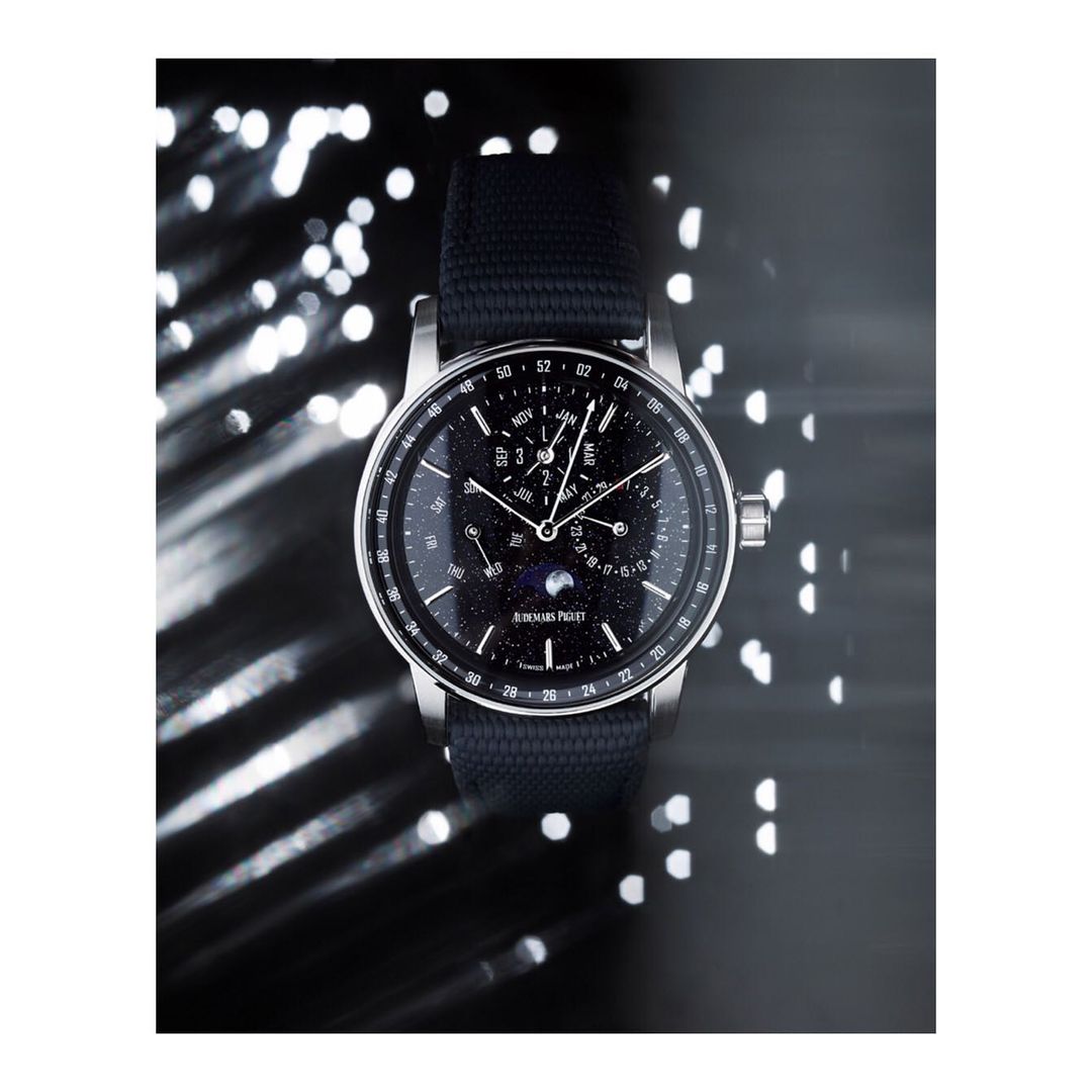 Luxury wristwatch with a dark band and black dial, highlighted by a silver casing and hands, set against a bokeh background.