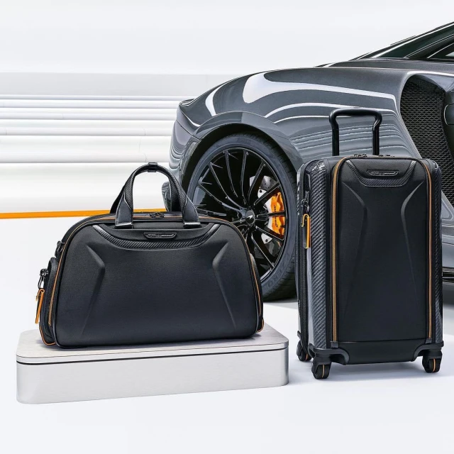 Black McLaren duffel and roller bag set on a plinth, with a McLaren car visible in the background, against a white backdrop.