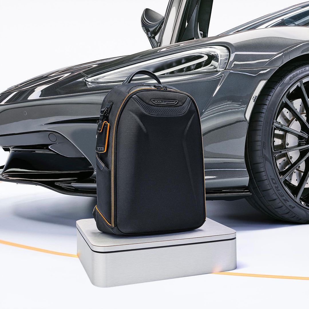 Black backpack sits on a white platform with a gray sports car in the background featuring a black tire, set in a studio environment.