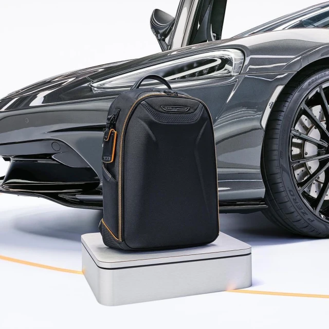 Black backpack sits on a white platform with a gray sports car in the background featuring a black tire, set in a studio environment.