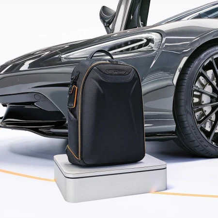 Black backpack sits on a white platform with a gray sports car in the background featuring a black tire, set in a studio environment.