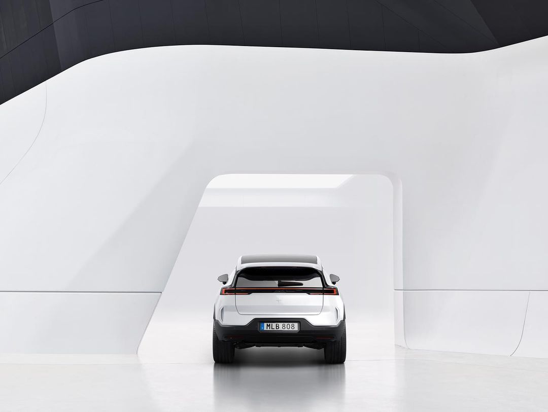 Rear view of a white Polestar SUV in a bright white architectural setting with rounded walls and indirect lighting, reflecting off the floor.