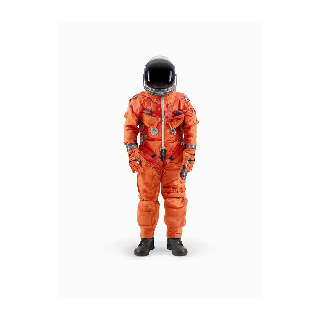 A person stands in a bright orange spacesuit with a helmet against a white backdrop, facing forward in a studio setting.