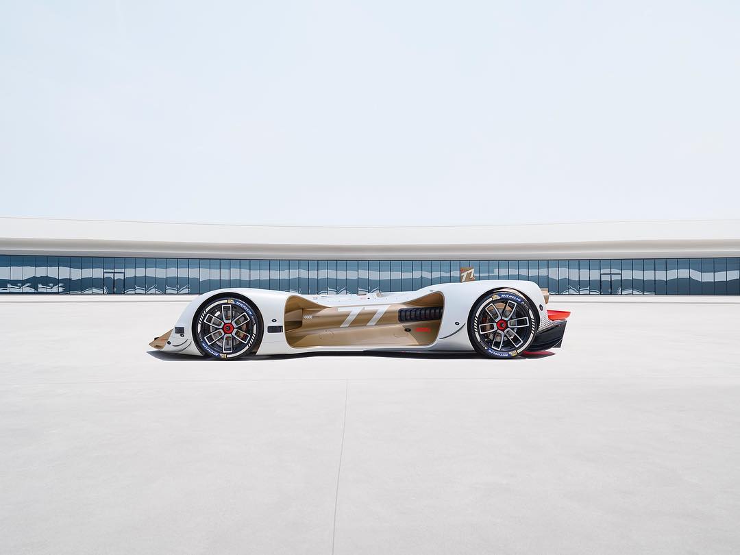 Side view of a futuristic white race car with red and black wheels on a smooth white surface with a modern building backdrop.
